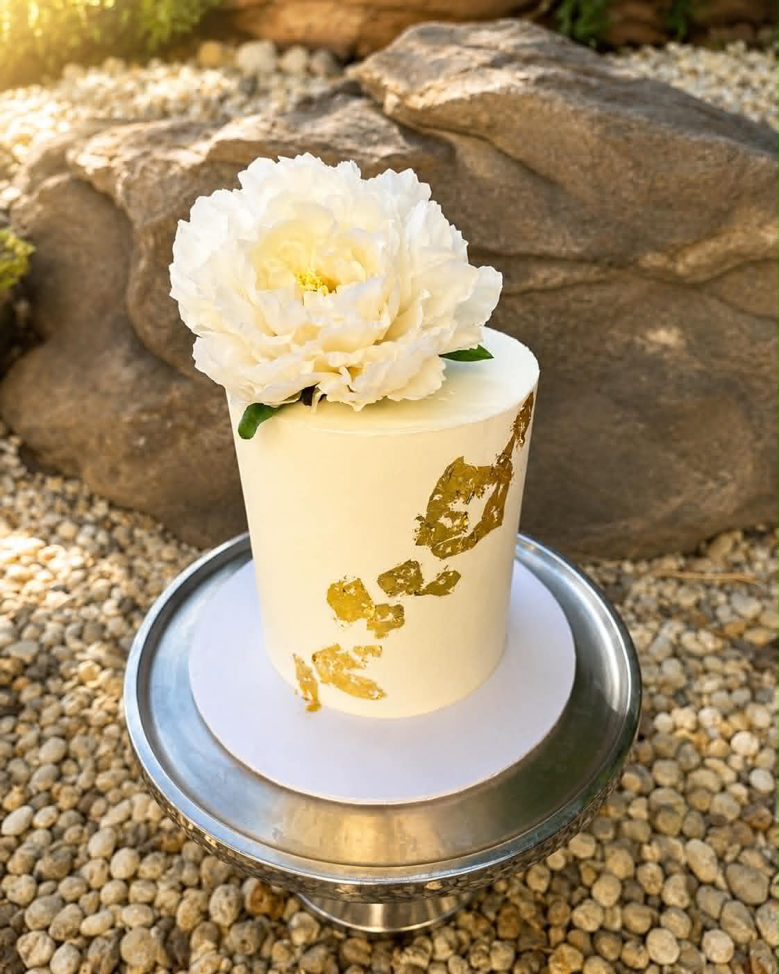Single tier white buttercream wedding cake with gold leaf accents and a large fresh white peony on top, displayed on a silver cake stand outdoors on pebbles